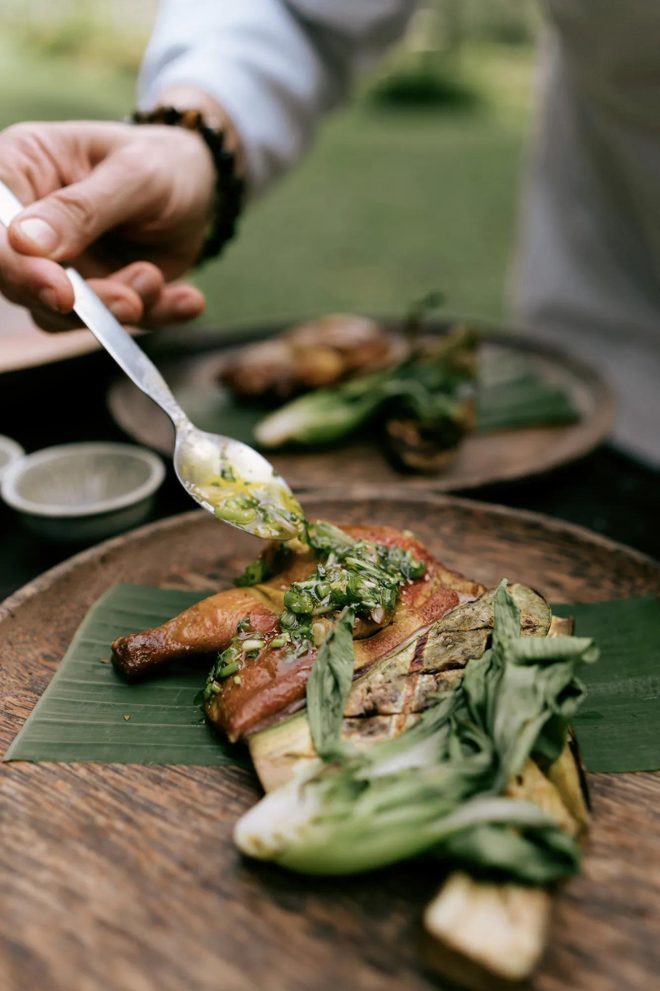 Grilled meat with herb sauce being spooned onto a wooden plate with vegetables.